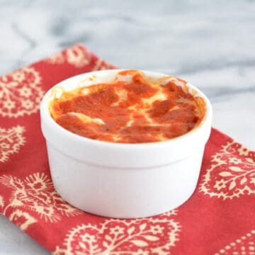 Ricotta cheese baked in a white ramekin topped with pasta sauce on a white table with a red napkin.